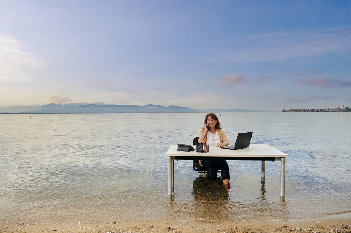Ein Schreibtisch steht im Bodensee. An ihm sitzt eine Frau, die ihre Füße im See abkühlt und dabei telefoniert. Ganz nach dem Motto "Arbeiten wo andere Urlaub machen".